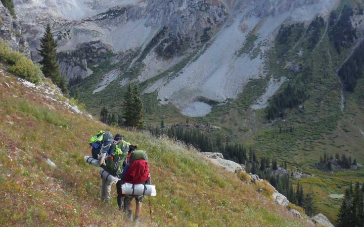 a group of students wearing backpacks hike across a grass field high above a mountainous valley